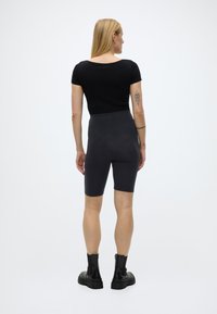 Femeie cu păr blond, purtând un top negru cu mâneci scurte, pantaloni scurți de ciclism gri închis, cizme de piele negre și tatuaje pe brațe, stând cu spatele spre camera.