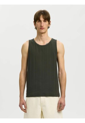 Hombre con una camiseta sin mangas acanalada de color verde oscuro y pantalones beige claro, de pie sobre un fondo blanco liso.