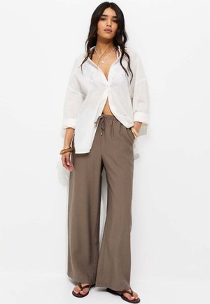 Frau trägt lockeres weißes Hemd mit Knöpfen, braune weite Hose mit Kordelzug, Sandalen und geschichtete Halsketten, steht vor weißem Hintergrund.