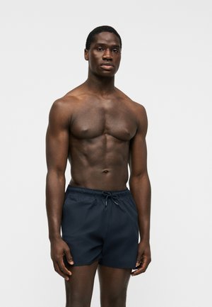 Uomo muscoloso in piedi, indossa pantaloncini da bagno blu scuro con coulisse, su uno sfondo grigio chiaro uniforme, braccia rilassate lungo i fianchi.