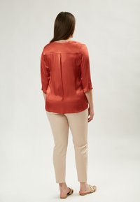 Seidenartige rostfarbene Bluse mit dreiviertelärmeln, Rückenschlitzausschnitt und glatter Textur, kombiniert mit beigen, eng anliegenden Hosen und Sandalen.