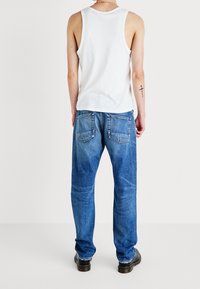 Man som bär blå jeans, svarta skor och en vit ärmlös topp, stående med ryggen mot kameran mot en enkel vit bakgrund.