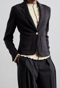 Černý přizpůsobený blejzr s dvěma předními kapsami a zapínáním na jeden knoflík; spárovaný se krémovým topem s vlnitým černým lemem u výstřihu.