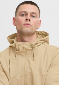 Beige zippered jacket with a hood, featuring drawstrings and a smooth texture; appears lightweight and casual, framed closely to the face.