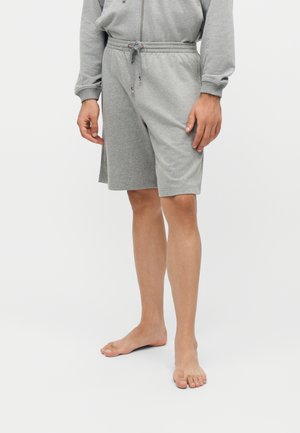 Personne portant un short gris à cordon et un haut gris assorti zippé, debout pieds nus sur un fond blanc uni.