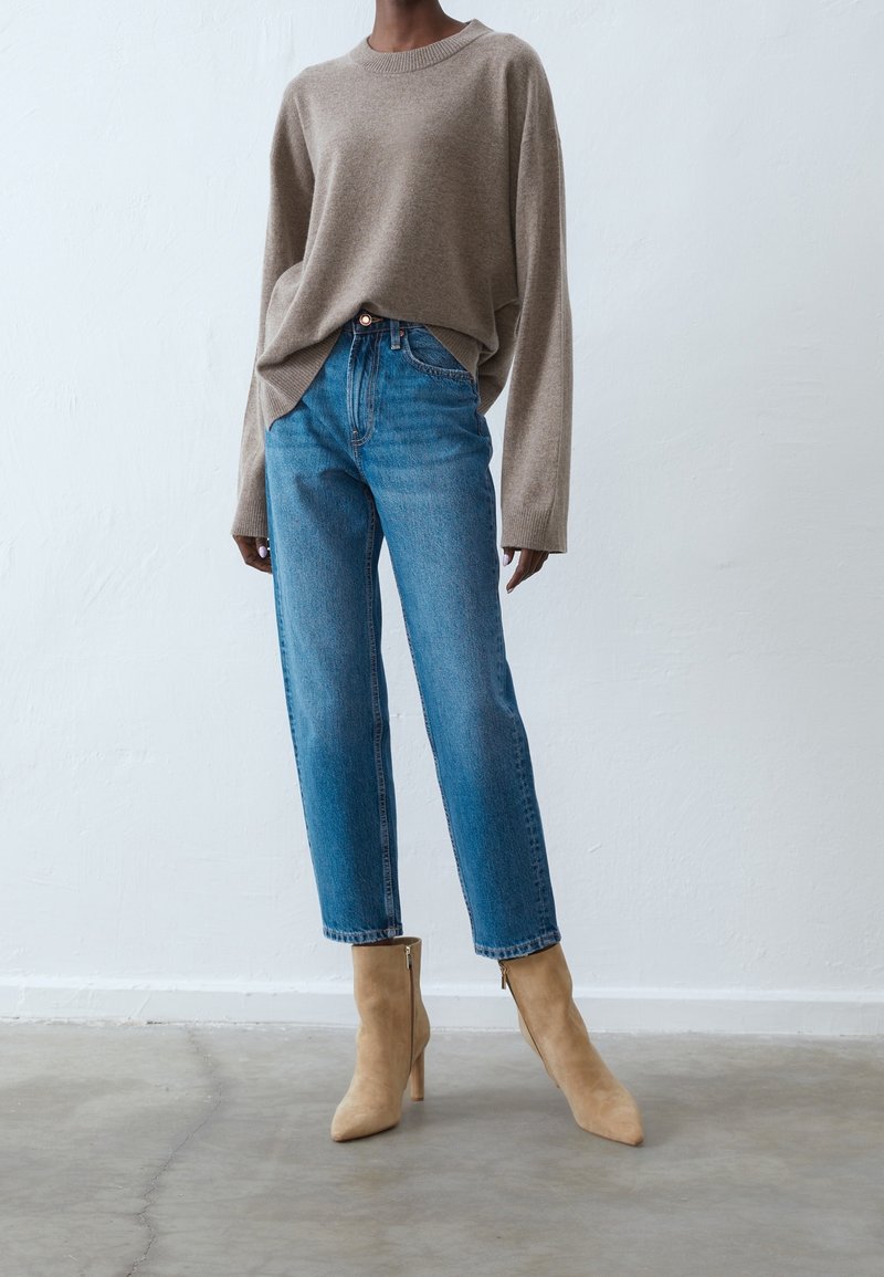 Personne portant un pull beige ample, un jean taille haute bleu et des bottines en daim marron clair, debout sur un sol en béton.