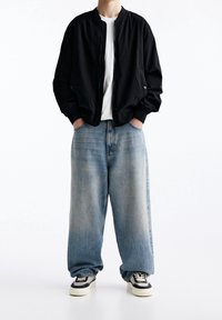 Zwarte bomberjack over een wit T-shirt, gecombineerd met lichtblauwe oversized jeans en zwart-witte sneakers. Eenvoudig ontwerp, relaxte pasvorm.