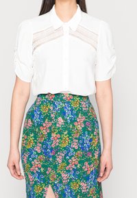 White short-sleeve blouse with a button-up front and lace panels; paired with a green floral print skirt featuring various colorful flowers.