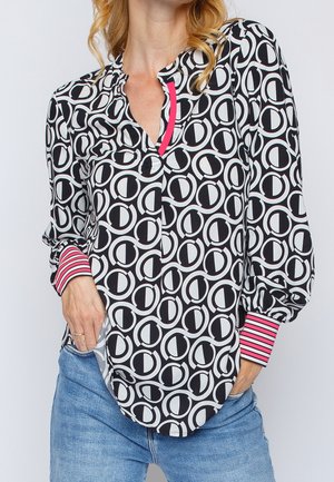 Mujer con blusa blanca y negra con estampado de diseños circulares, puños a rayas rosas y vaqueros azules, manos parcialmente en los bolsillos.