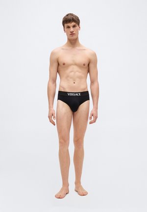 Noor mees paljajalu, kandes musti Versace aluspükse, seisab tavalise heleda halli tausta ees.