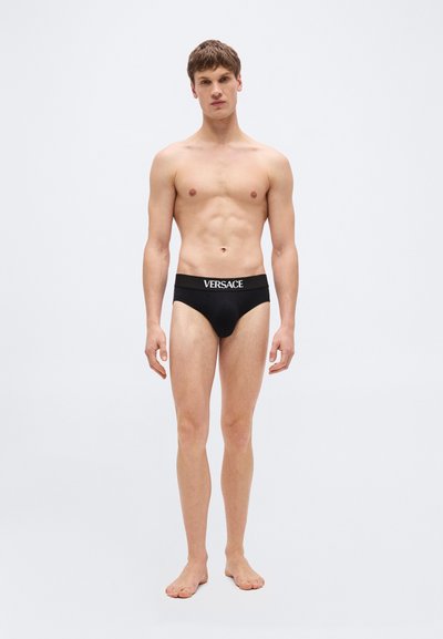 Jeune homme debout pieds nus portant un slip noir Versace sur un fond gris clair uni.