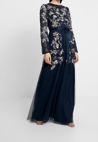 Lång marinblå klänning med långa genomskinliga ärmar, dekorerad med silverfärgade blommönster och pärlor; har ett smalnat midjeband och en flödande kjol.