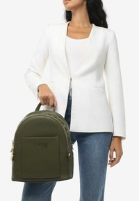 Zaino in pelle verde di forma rotonda, con logo impresso sulla parte anteriore, chiusura con zip e manici imbottiti. Modello che indossa un blazer bianco e jeans.
