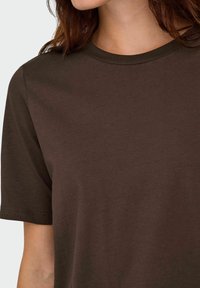 Primo piano di una persona che indossa una semplice t-shirt a maniche corte di colore marrone scuro, che mostra parte del collo e della spalla.