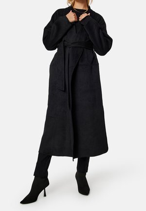 Kobieta w długim czarnym płaszczu z paskiem, czarnych spodniach i czarnych, zaostrzonych wysokich butach na obcasie, stojąca z rękami przy kołnierzu.