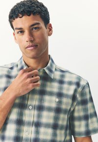 Short-sleeve plaid shirt in green, blue, and cream. Features a button-up collar, front buttons, and a single pocket on the chest.