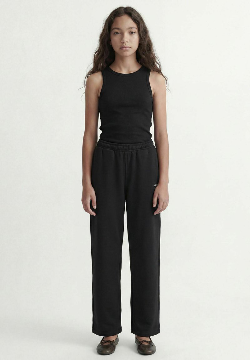 Jeune personne aux longs cheveux bouclés portant un haut noir sans manches, un pantalon noir ample et des ballerines noires, debout devant un fond uni.