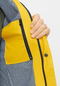 Gul og navy stribet regnjakke indvendigt med en lynlåslomme, glat tekstur og kontrasterende farvepaneldetaljer.