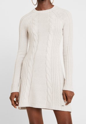 Frau in einem langen, cremefarbenen Strickkleid mit Kabelmuster und gerippten Bündchen und Saum, accessorized mit großen Creolen.