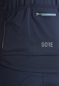 Marinblå idrottsklädsel med dragkedjeficka, platta sömmar och reflexdetaljer. Materialet verkar lättviktigt med en slät yta.