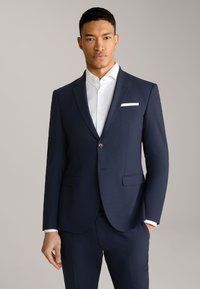 Giacca da abito blu in tessuto testurizzato con revers a punta, chiusura con un solo bottone e un fazzoletto bianco in tasca come dettaglio.