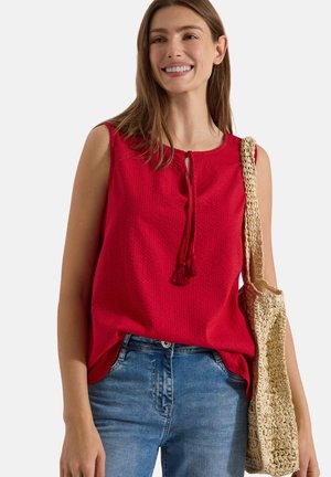 Femme souriante portant un haut rouge sans manches, un jean bleu et un sac en paille tressée sur l'épaule, debout devant un fond uni.