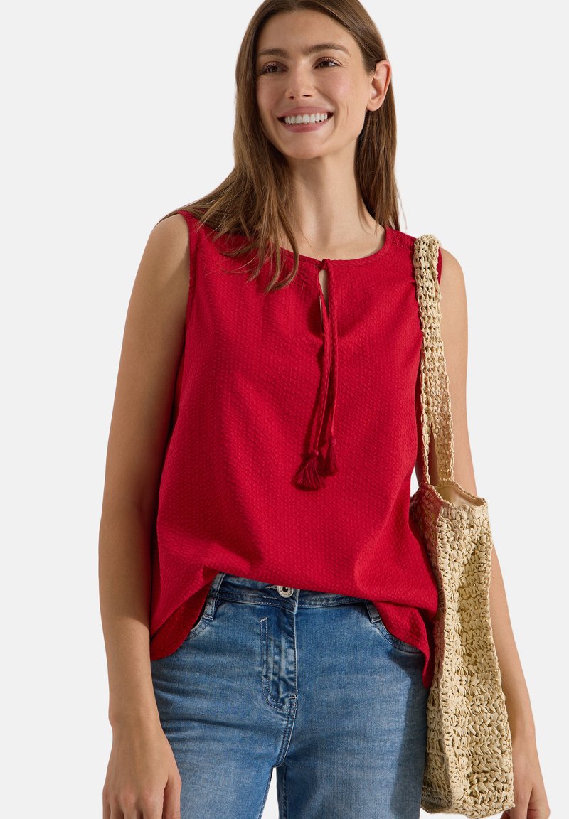 Femme souriante portant un haut rouge sans manches, un jean bleu et un sac en paille tressée sur l'épaule, debout devant un fond uni.