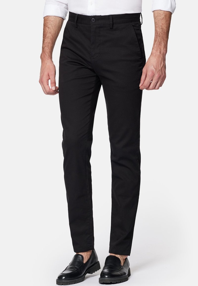Homme portant un pantalon noir slim, une chemise blanche aux manches retroussées et des mocassins noirs en cuir, debout devant un fond uni.
