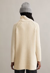 Cremefarbenes Strickpullover mit hohem Kragen, strukturiertem Muster und lockerer Passform. Der Rücken hat Raglanärmel und seitliche Schlitze.