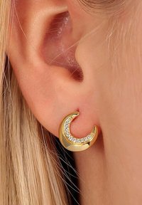 Gula halvmåneformade örhängen med inlagda klara rhinestones, med en polerad slät yta och en böjd design, bärs på örsnibben.