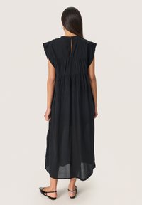 Schwarzes Midi-Kleid mit kurzen, flatternden Ärmeln, gerafftem Taillenbereich und Rückendetail mit Schlitz. Aus leichtem, strukturiertem Stoff gefertigt. Flache Sandalen getragen.
