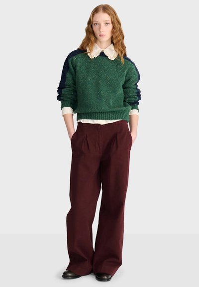 Jeune femme aux cheveux roux porte un pull vert et bleu marine par-dessus une chemise blanche à col, un pantalon large bordeaux et des chaussures noires.