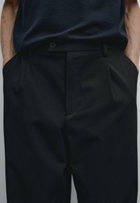 Black trousers made of smooth fabric, featuring a tailored fit, central crease, and side pockets. Waistband includes a button closure.