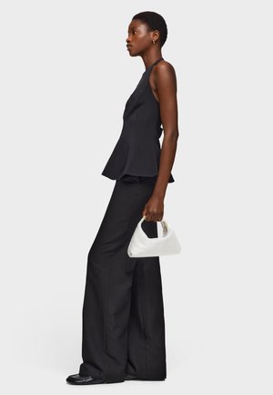 Mujer con top negro sin mangas y pantalones negros de pierna ancha, sosteniendo un pequeño bolso blanco, de perfil contra un fondo blanco.