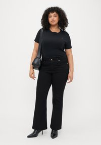 T-shirt noir à manches courtes, jean noir taille haute, escarpins noirs à talons pointus et un petit sac à main noir. Cheveux bouclés et fond neutre.