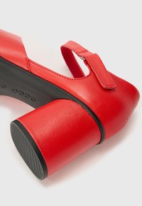 Red leather pump with a thick black block heel, featuring a round toe and a loop strap across the foot with a metallic buckle.