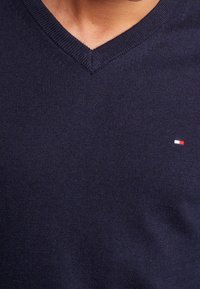 Ciemnoniebieski sweter w serek z gładkiej dzianiny, z małym czerwono-białoniebieskim logo po lewej stronie klatki piersiowej.