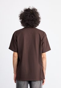 T-shirt in cotone marrone con vestibilità rilassata e maniche corte. Caratterizzato da un colletto a girocollo e un motivo a tinta unita, indossato con jeans grigi.