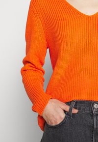 Oranger Strickpullover mit V-Ausschnitt, langen Ärmeln und texturiertem Muster, kombiniert mit dunkelgrauen High-Waist-Jeans.