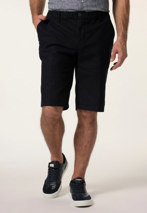 Mann trägt schwarze knielange Shorts, graues Shirt und schwarze Sneaker, geht mit einer Hand in der Tasche vor hellem Hintergrund.