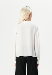 Blusa bianca a maniche lunghe con una vestibilità rilassata, tessuto morbido, maniche arricciate e dettaglio con bottoni ai polsini. L'orlo arriva leggermente sotto la vita.