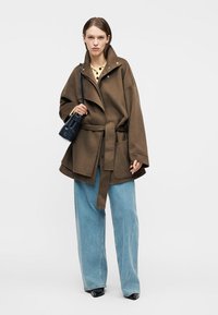 Femeie purtând o haină maro supradimensionată cu curea, blugi largi bleu deschis, pantofi negri cu vârf ascuțit, top galben și o geantă mică neagră de umăr.