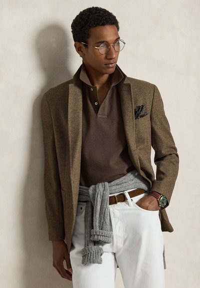 Mees kandmas ümmarguseid prille, pruunikat bleiserit, pruuni särki, valgeid pükse, vöökohal seotud halli kampsunit ja käekella, seisab heleda seina ääres.