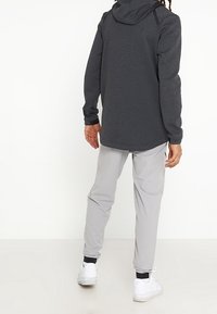 Veste à capuche grise avec des accents noirs, assortie à un pantalon gris clair et des baskets blanches. Le tissu semble à la fois lisse et texturé.