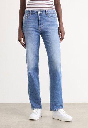 Person iført lyseblå jeans med lige ben, hvide sneakers og en stribet top, stående på en lys gulvbelægning mod en ensfarvet hvid væg.