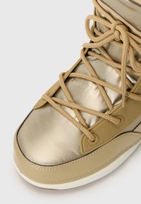 Gros plan d'une sneaker dorée avec un laçage complexe et des détails texturés sur le côté, sur un fond uni.