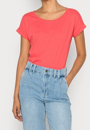Kvinde iført en koralfarvet t-shirt med korte ærmer stoppet ned i højtsiddende lyseblå jeans med hænderne afslappede, den ene i lommen.