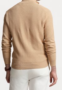 Homme portant un pull en maille beige texturée et un pantalon blanc, vu de dos avec une montre marron au poignet gauche.