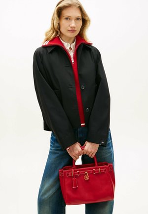 Joven mujer con chaqueta negra sobre un suéter rojo con cremallera, sosteniendo un bolso rojo con detalles dorados frente a ella.