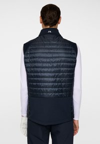 Marinblå pufferväst med kviltad textur, hög krage och logodetalj på baksidan. Har ett mjukt material och en åtsittande design för värme.